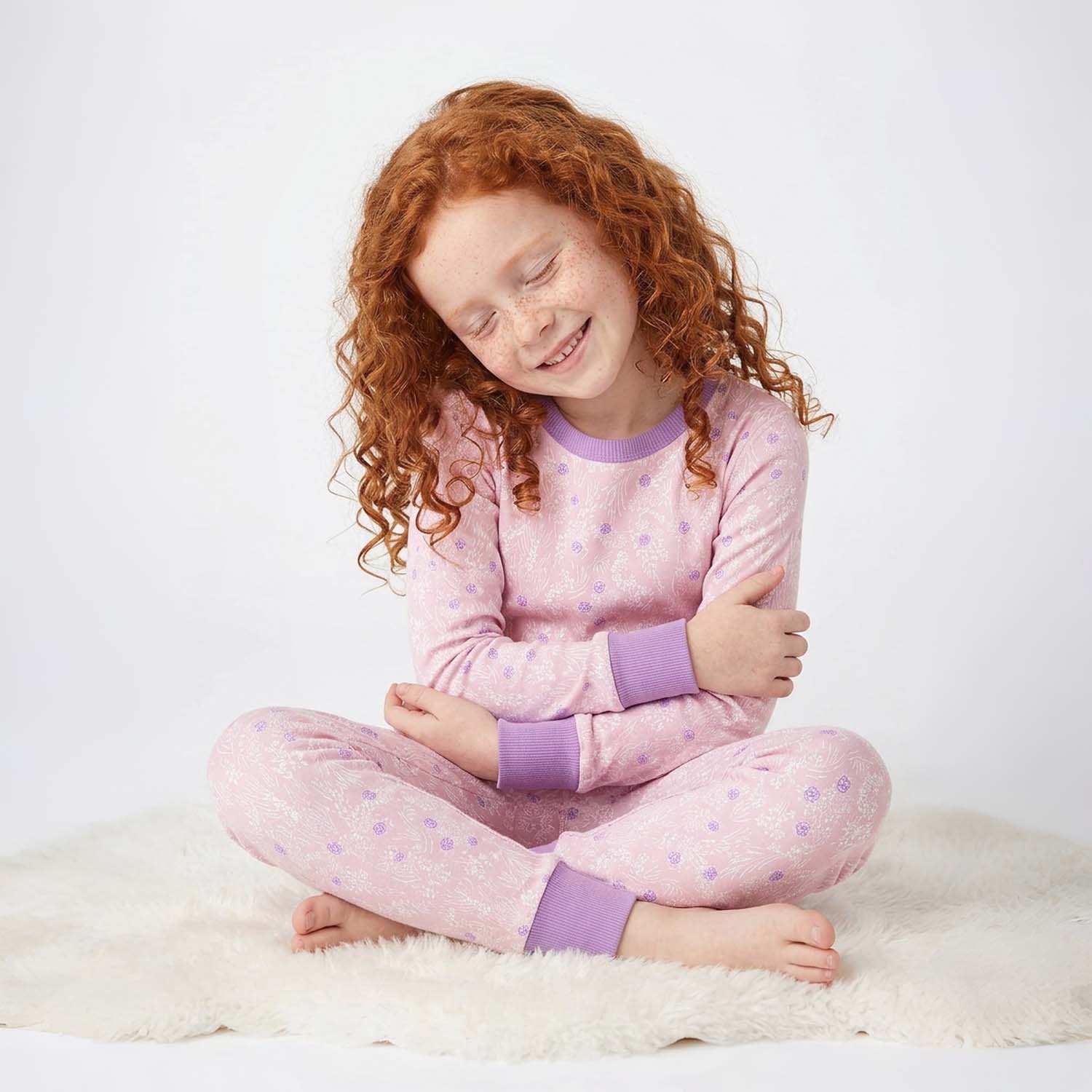 Child wearing pink pajamas with purple cuffs and trim sitting on a white surface.