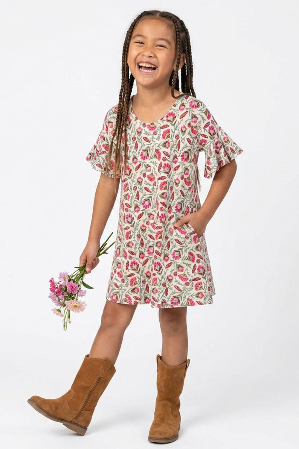 A young girl with braided hair is smiling and laughing while wearing a short-sleeve dress with a pink and green floral pattern on a cream background. She is holding a small bouquet of pink flowers and wearing brown suede boots, standing against a solid white studio background.