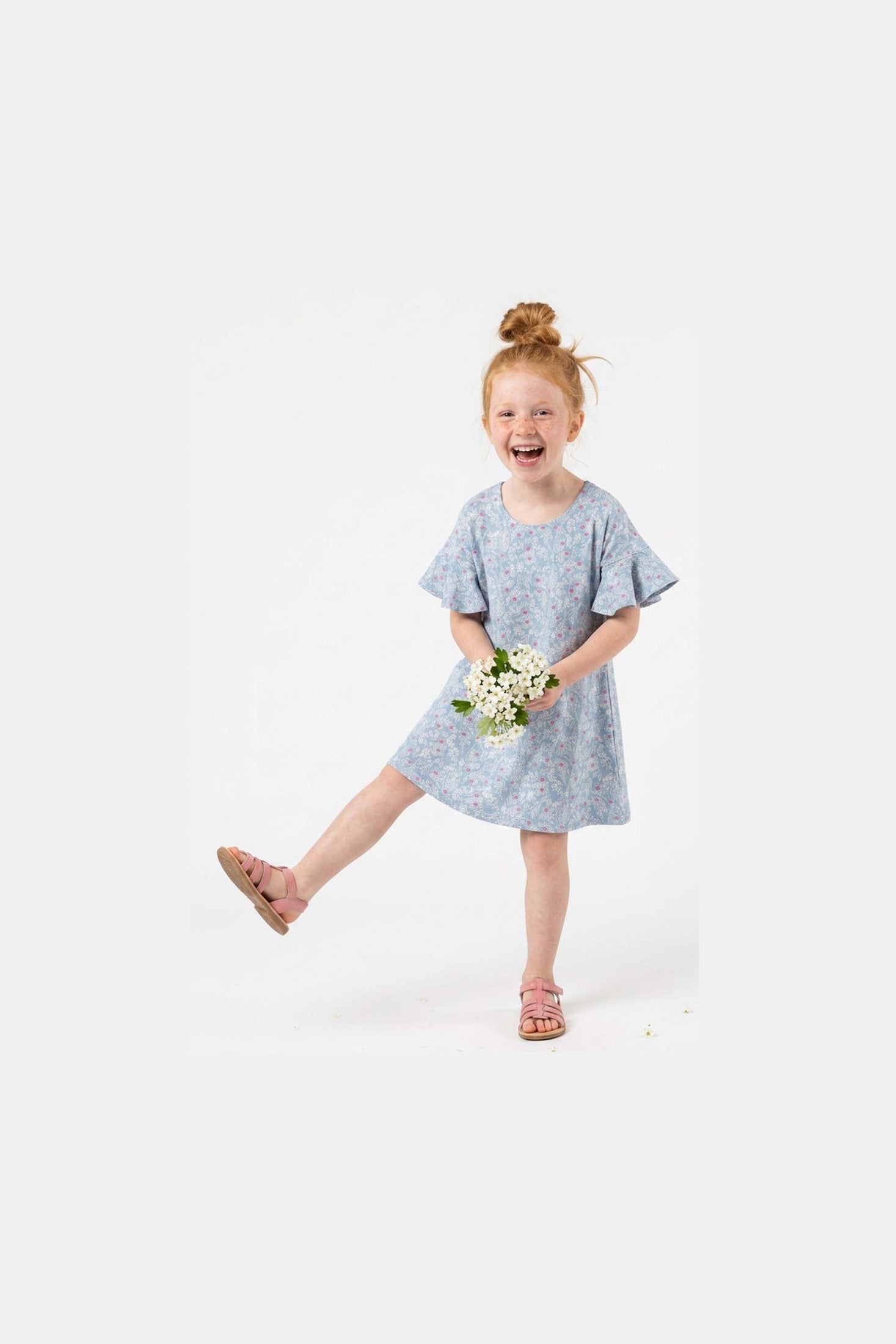 A young girl with red hair in a bun is wearing a light blue short-sleeve dress with a small floral print and holding a bouquet of white flowers. She is wearing pink sandals and posing on a solid white studio background.