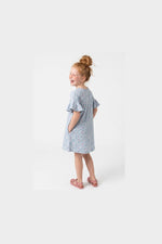 A young girl with red hair in a bun is shown from the back wearing a light blue short-sleeve dress with a small pink and green floral pattern. She is standing against a clean white studio background.