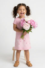 A young girl with curly brown hair in pigtails stands smiling and holding a bouquet of pink and white peonies. She is wearing a light pink short-sleeve dress with white floral embroidery and tan sandals, standing next to a small wooden stool against a white studio background.