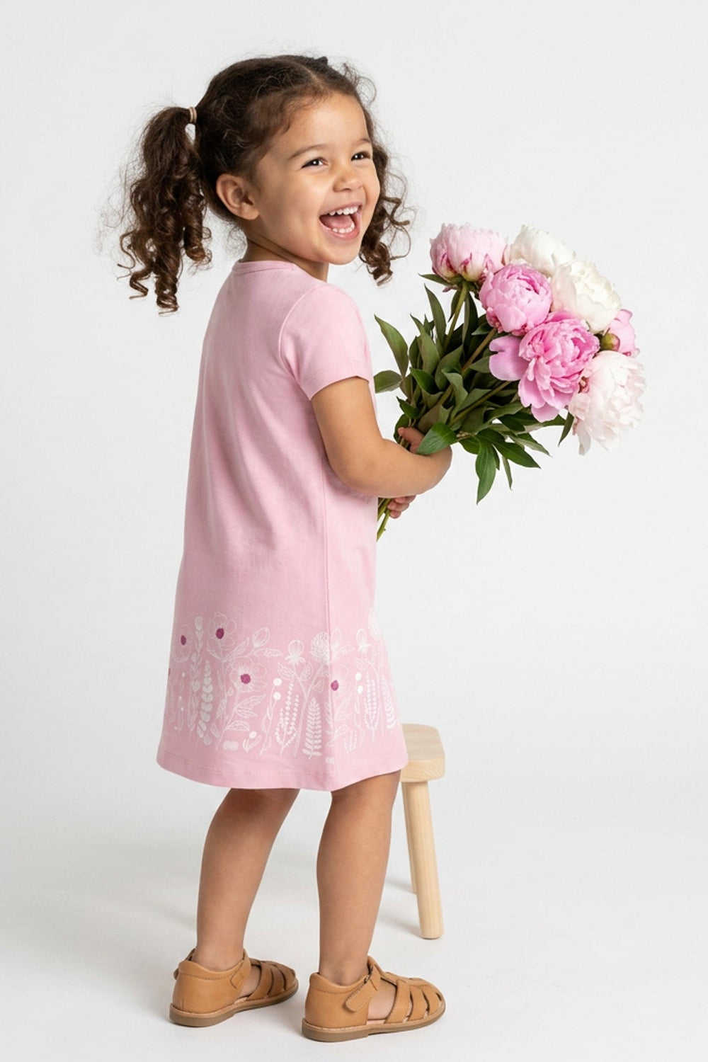 A young girl with curly brown hair in pigtails stands smiling and holding a bouquet of pink and white peonies. She is wearing a light pink short-sleeve dress with white floral embroidery near the hem, paired with tan leather sandals, and is posed next to a small wooden stool against a plain white studio background.