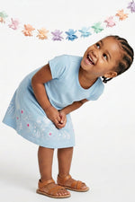 A young child wearing a light blue short-sleeve dress with white floral embroidery and brown leather sandals, standing against a white studio background under a pastel flower garland.
