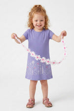 A young girl with curly red hair stands against a light gray studio background, holding up a pink and white daisy flower garland. She is wearing a lavender purple short-sleeve dress with a floral print near the hem and pink sandals.