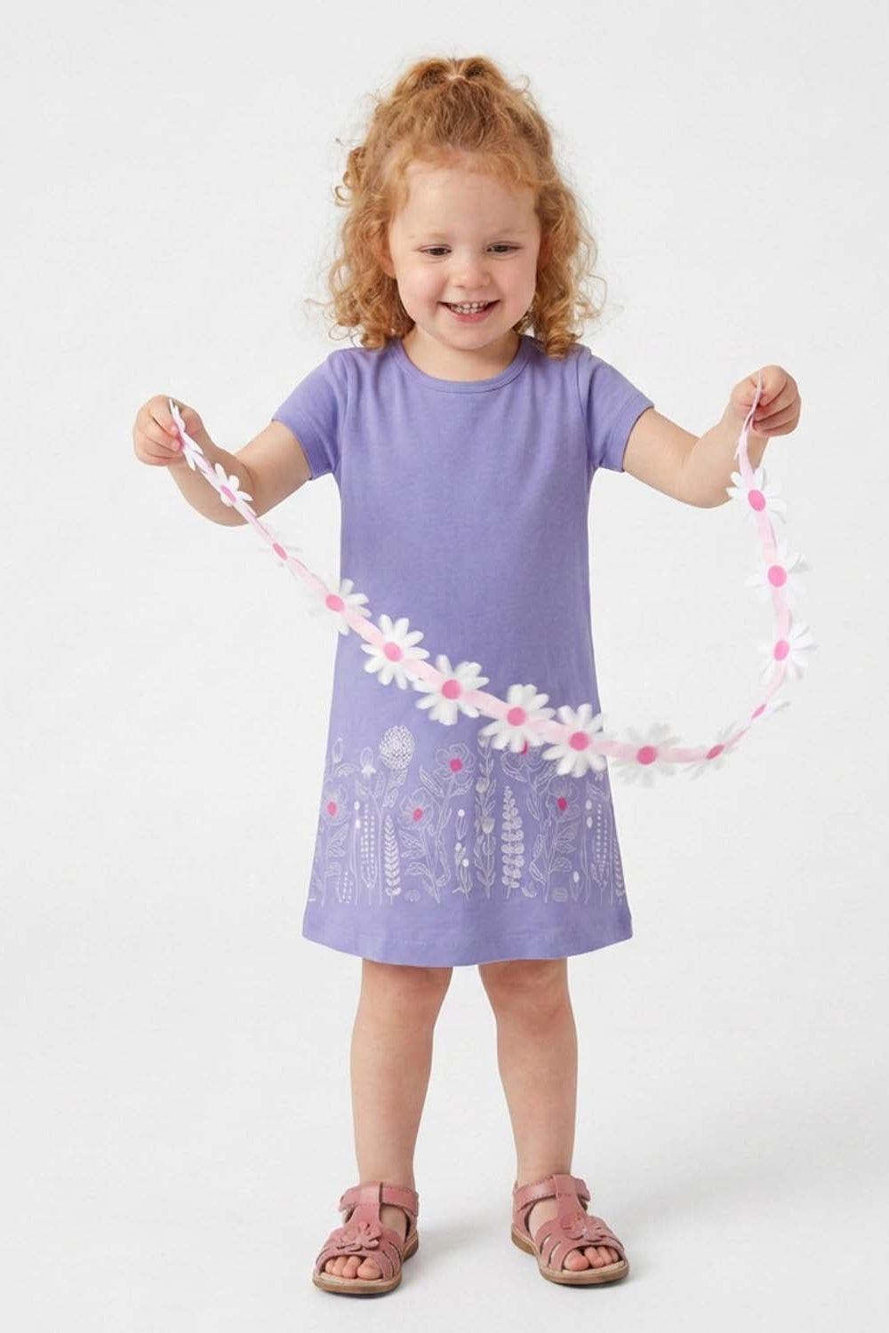 A young girl with curly red hair stands against a light gray studio background, holding up a pink and white daisy flower garland. She is wearing a lavender purple short-sleeve dress with a floral print near the hem and pink sandals.