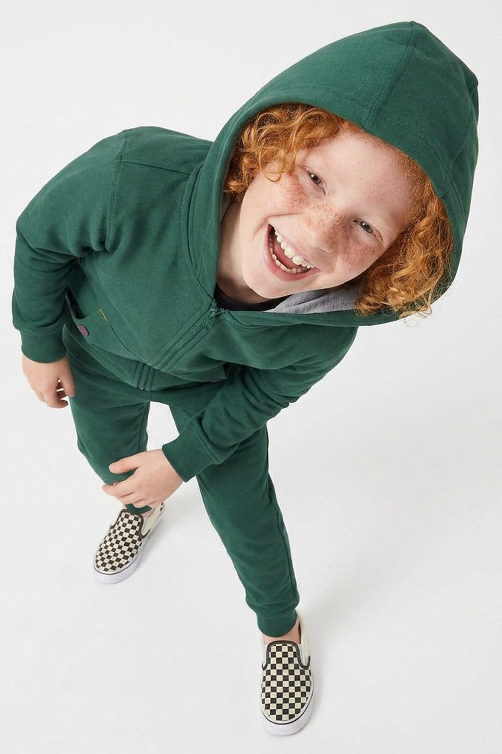 A smiling child with curly red hair wearing a matching dark green hooded sweatshirt and jogger pants, paired with black-and-white checkered slip-on shoes, photographed against a clean white studio background.