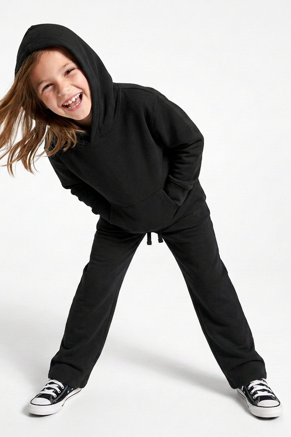 A young girl wearing a matching black hooded sweatshirt and sweatpants set, posing dynamically on a clean white studio background.
