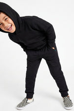 A young boy wearing a matching black hooded sweatshirt and jogger pants set, paired with black and white checkered slip-on sneakers, posing against a solid white studio background.