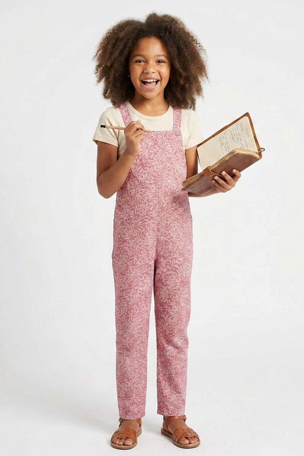 A young girl with curly brown hair stands against a white studio background, wearing pink and white floral print overalls over a cream t-shirt and brown sandals. She holds an open book and a pencil.