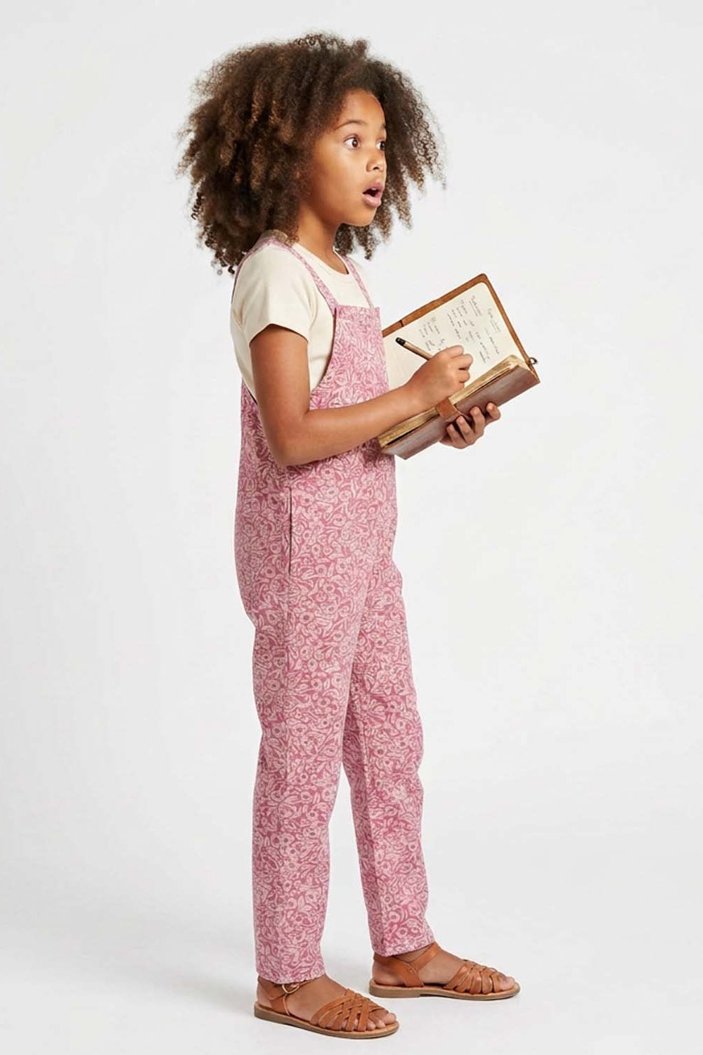 A young girl with curly brown hair stands in profile wearing pink floral overall pajamas over a white t-shirt, holding a notebook and pen against a plain white studio background.
