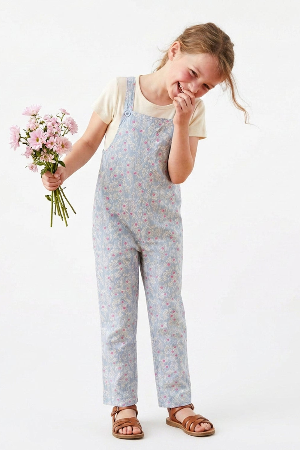 A young girl wearing a light blue floral print overall jumpsuit over a cream t-shirt and brown sandals, holding a bouquet of pink flowers against a plain white background.