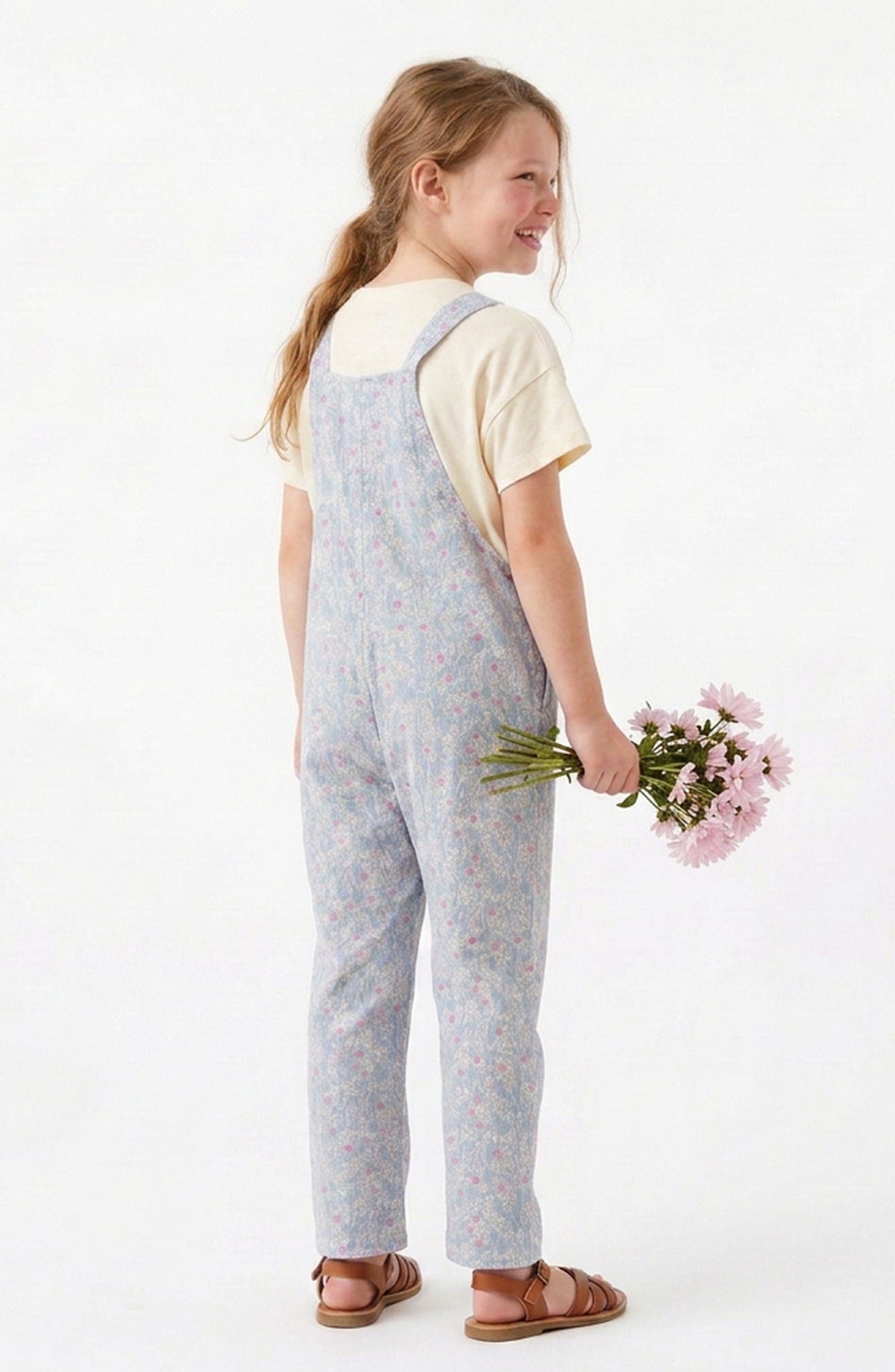 A young girl with blonde hair in a ponytail is shown from the back, wearing light blue floral print overall over a cream t-shirt and brown sandals, holding a small bouquet of pink flowers against a plain white background.