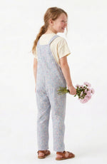 A young girl with blonde hair in a ponytail is shown from the back, wearing light blue floral print overall over a cream t-shirt and brown sandals, holding a small bouquet of pink flowers against a plain white background.