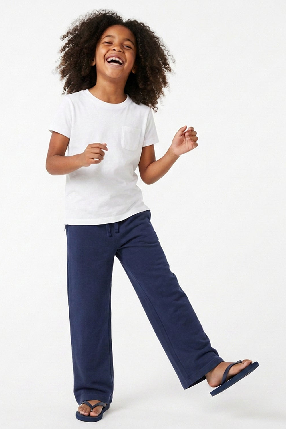 A young girl with curly dark hair is smiling and laughing while posing in a white short-sleeve crewneck t-shirt, navy blue wide-leg pants, and dark blue flip-flops against a solid white studio background.
