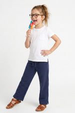 A young girl with glasses is modeling a white short-sleeve pocket t-shirt and navy blue pants, holding a rainbow-colored popsicle against a plain white studio background.