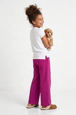 A young girl with her hair in a bun stands in profile, holding a small fluffy dog. She is wearing bright magenta wide-leg pants, a white t-shirt, and tan slippers against a clean white studio background.