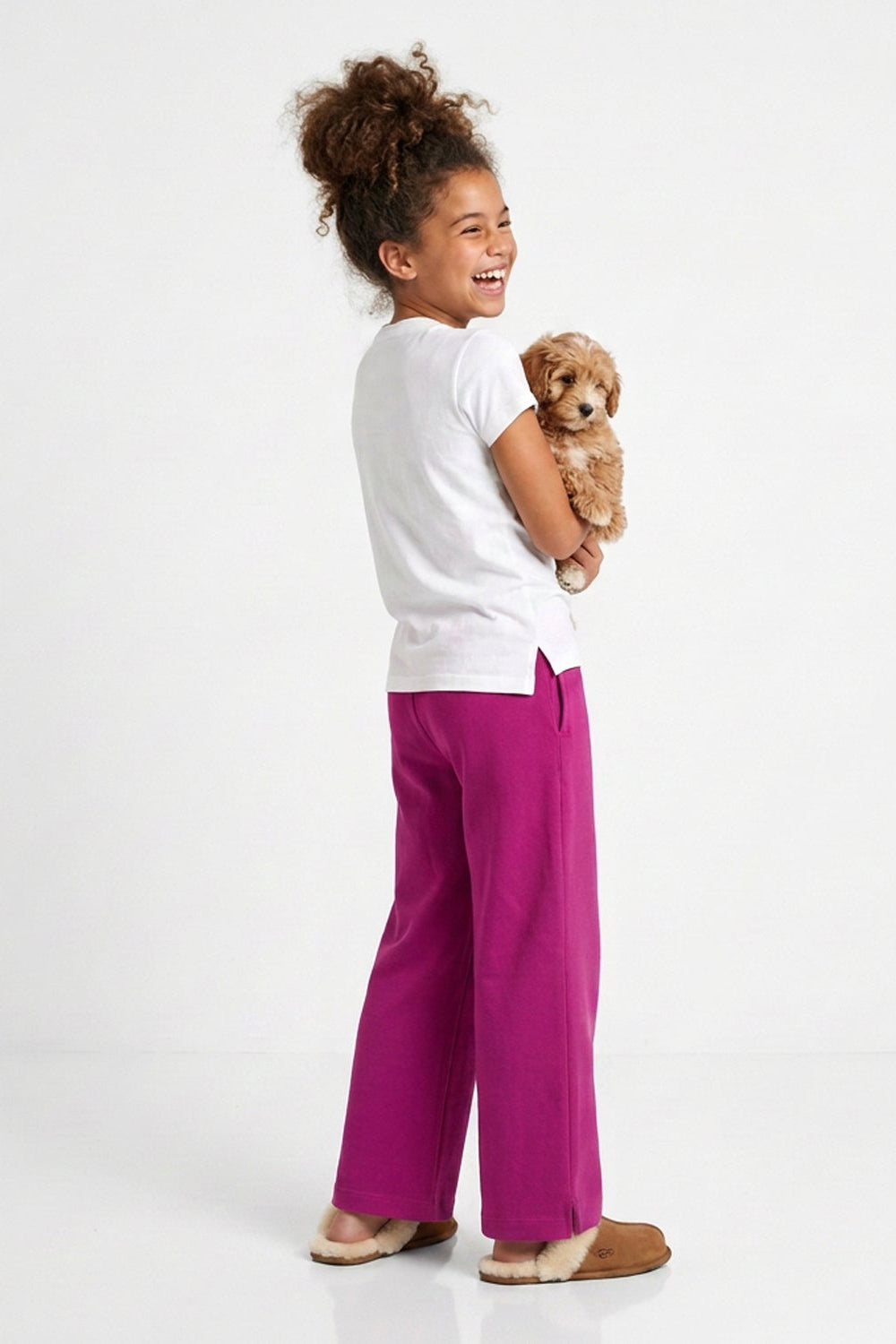 A young girl with her hair in a bun stands in profile, holding a small fluffy dog. She is wearing bright magenta wide-leg pants, a white t-shirt, and tan slippers against a clean white studio background.