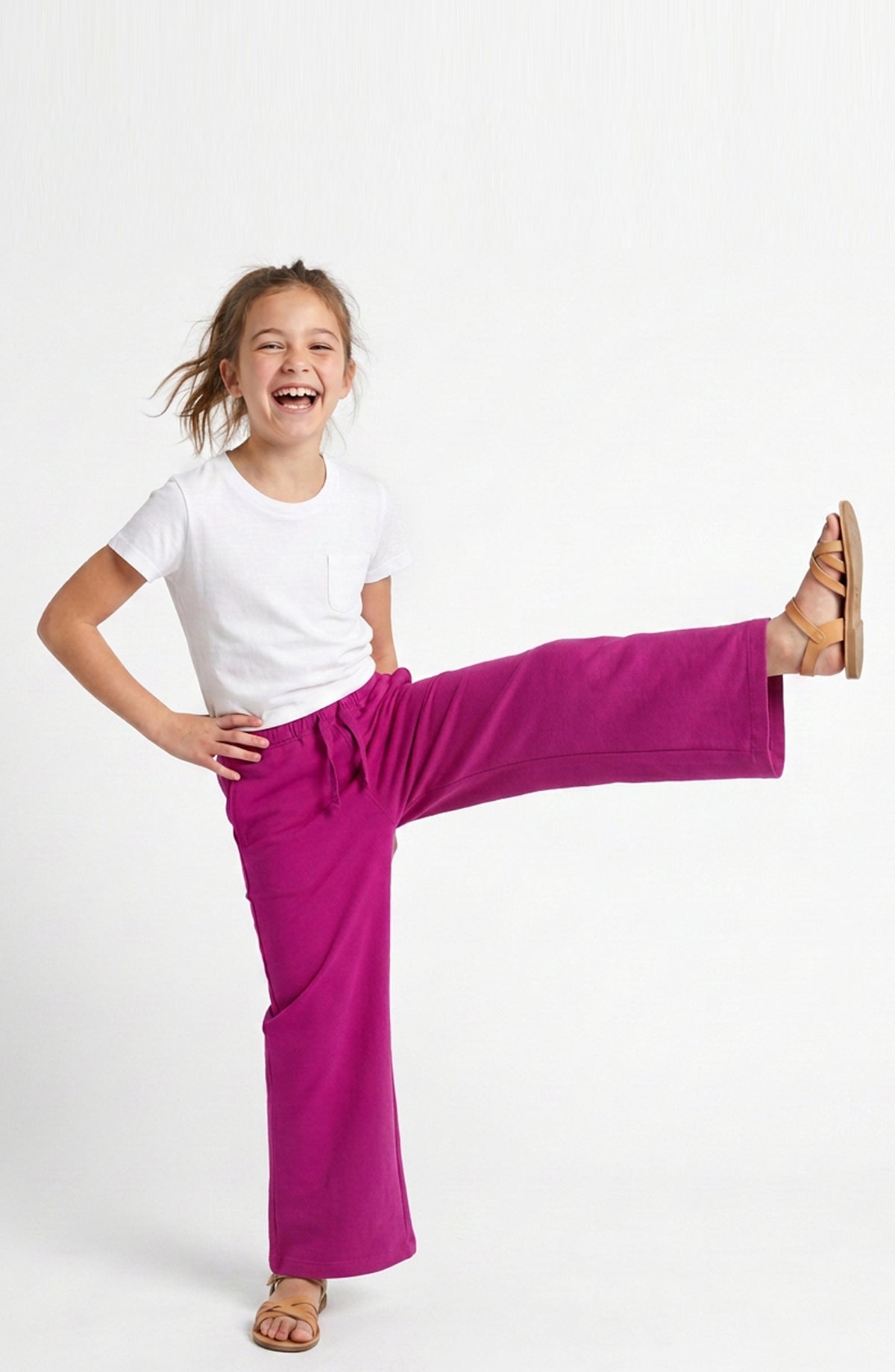 A young girl with blonde hair tied back, wearing a white t-shirt and bright magenta wide-leg pants, posing with one leg raised in a playful kick pose against a clean white studio background.