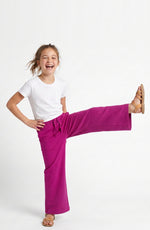 A young girl with blonde hair tied back, wearing a white t-shirt and bright magenta wide-leg pants, posing with one leg raised in a playful kick pose against a clean white studio background.