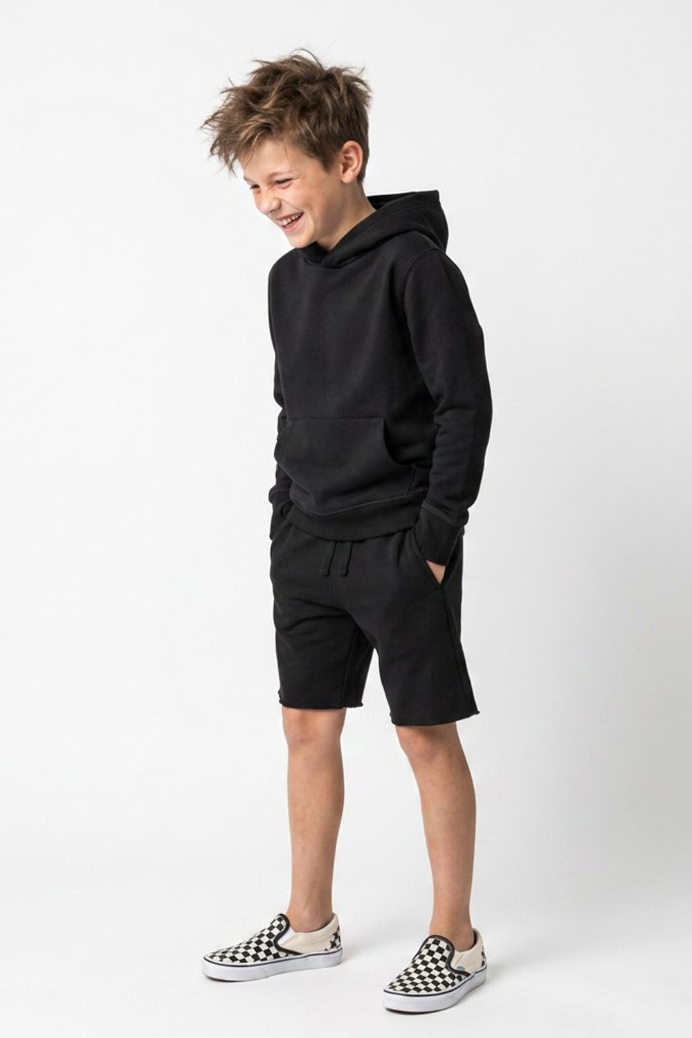 A young boy modeling a black two-piece loungewear set consisting of a pullover hoodie and matching drawstring shorts, paired with black and white checkered slip-on shoes, against a plain white studio background.