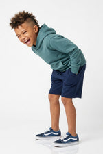 A young boy laughing and leaning forward, wearing a teal-green hoodie, navy blue shorts, and navy blue Vans sneakers with white stripes, photographed against a seamless white studio background.