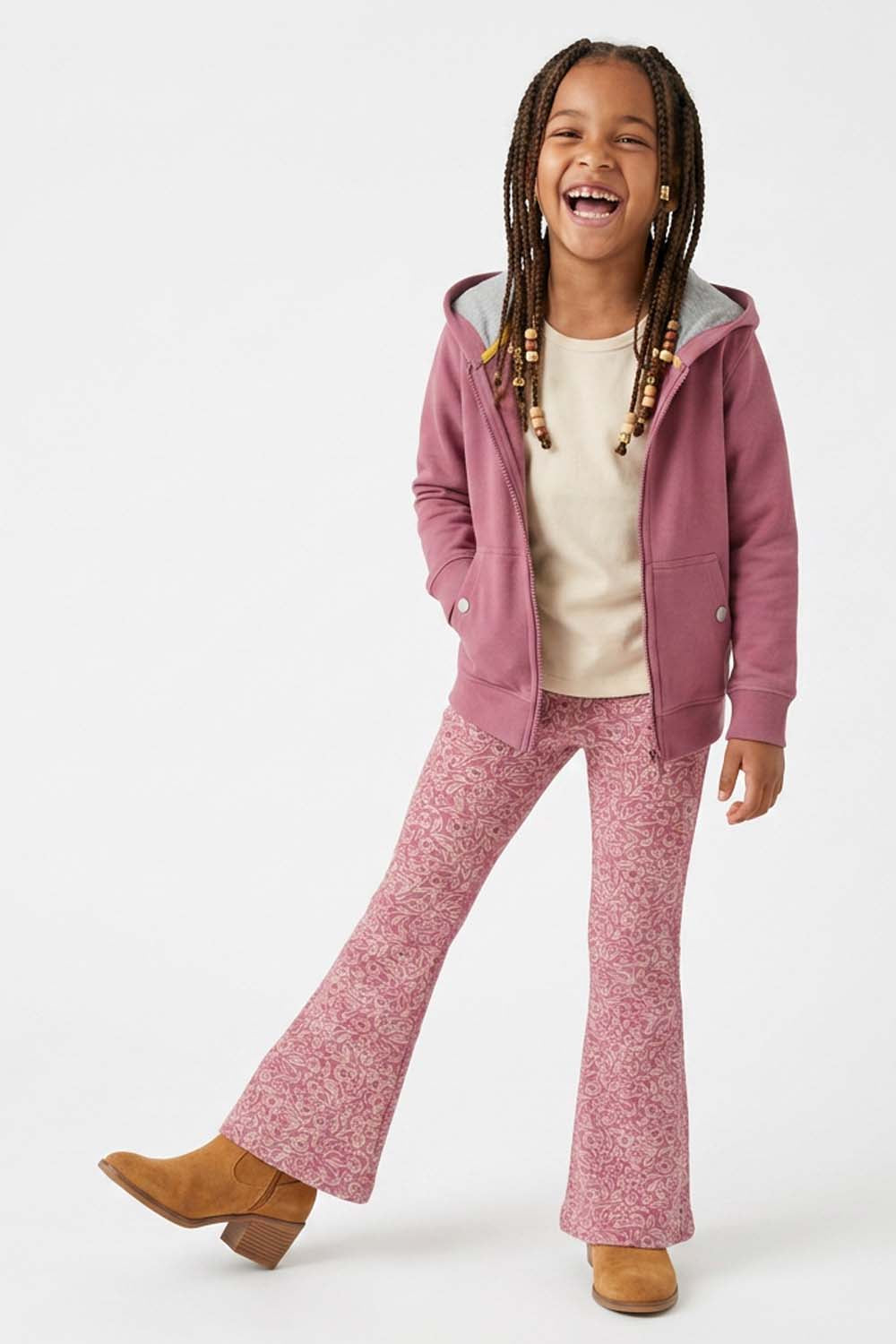 A young girl with braided hair is smiling and posing in a pink zip-up hoodie, matching pink patterned flared pants, a cream t-shirt, and tan boots against a plain white studio background.