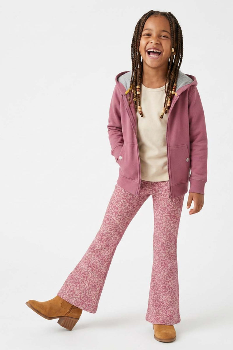A young girl with braided hair is smiling and posing in a pink zip-up hoodie, matching pink patterned flared pants, a cream t-shirt, and tan boots against a plain white studio background.