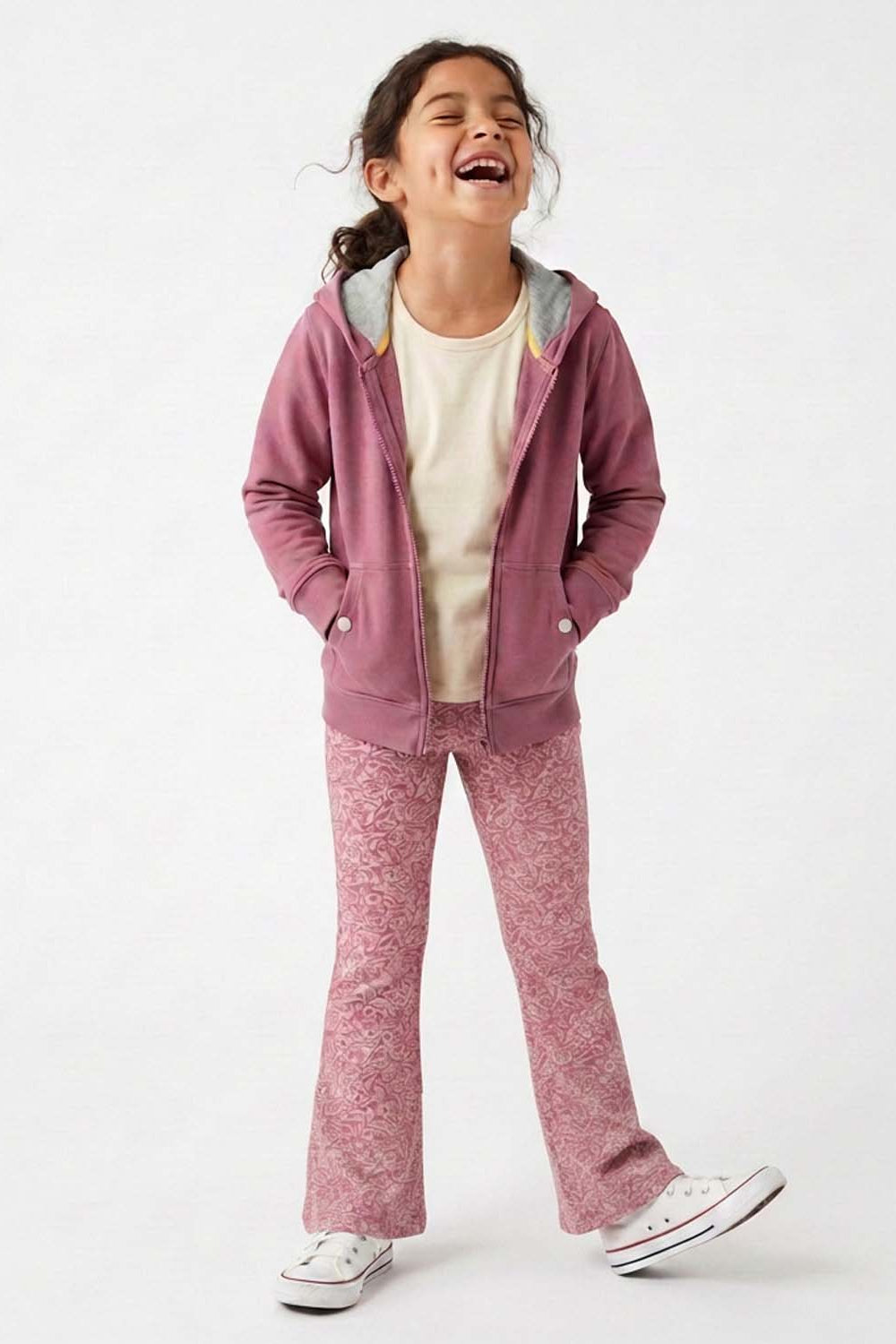 A young girl laughing while wearing a dusty rose zip-up hoodie, matching patterned pants, a cream t-shirt, and white sneakers against a plain white studio background.