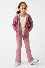 A young girl laughing while wearing a dusty rose zip-up hoodie, matching patterned pants, a cream t-shirt, and white sneakers against a plain white studio background.