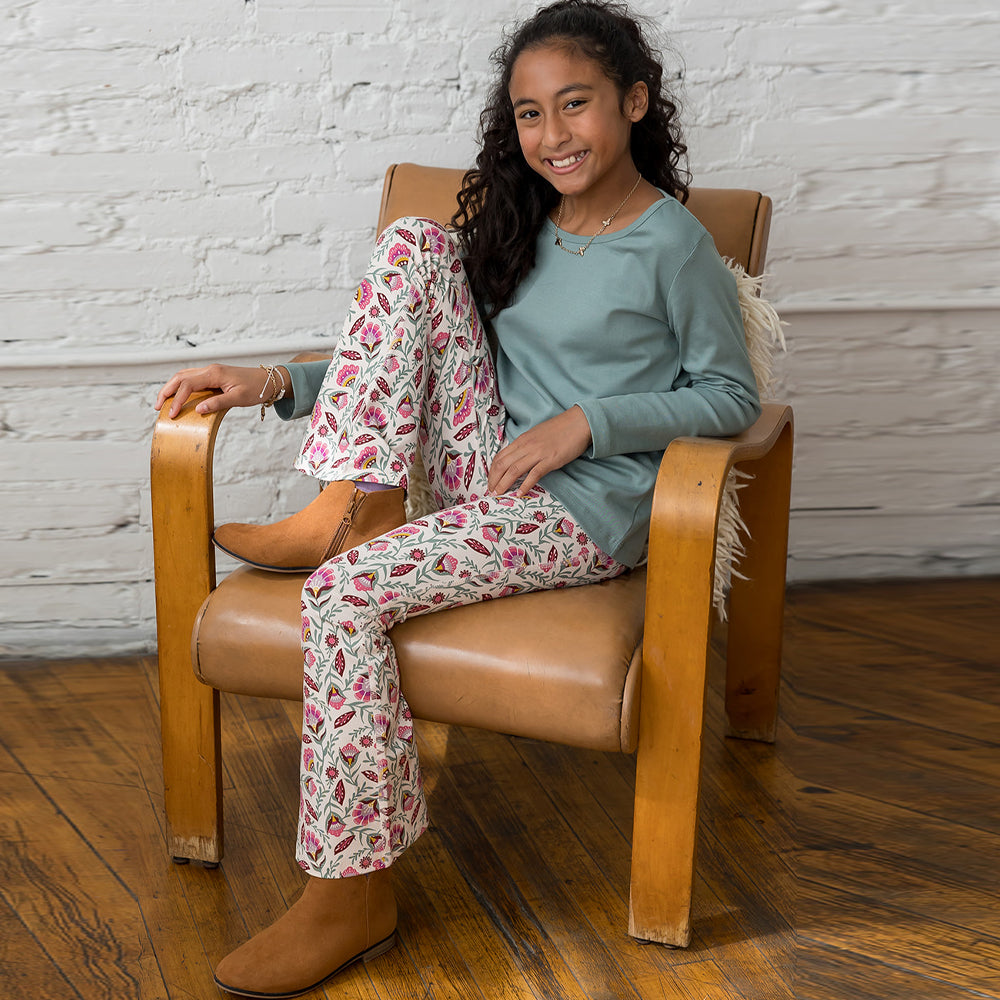 A young girl with curly dark hair sits smiling in a tan leather armchair with wooden arms, wearing a sage green long-sleeve top and cream pants with a pink and red floral pattern. She is wearing tan suede boots against a white brick wall and wooden floor background.