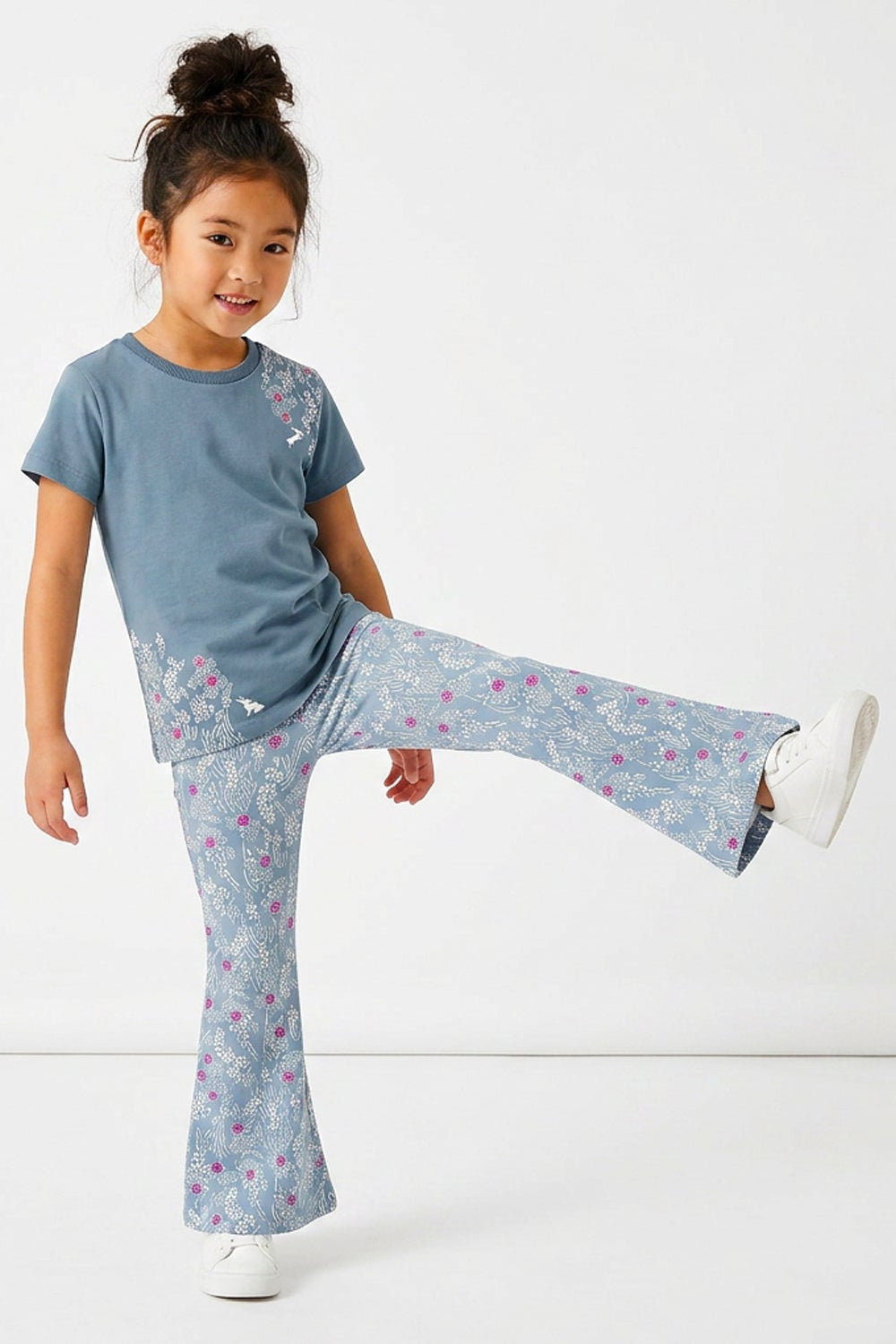 A young girl modeling a matching two-piece pajama set consisting of a slate blue short-sleeve t-shirt with a floral print and matching blue flared pants with a pink and white floral pattern, set against a plain white studio background.