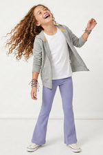 A young girl with long curly brown hair is captured mid-laugh, wearing a gray zip-up hoodie over a white t-shirt, lavender flare pants, and white sneakers. She is accessorized with colorful beaded bracelets against a clean white studio background.