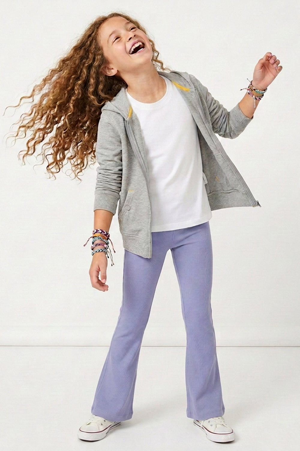 A young girl with long curly brown hair is captured mid-laugh, wearing a gray zip-up hoodie over a white t-shirt, lavender flare pants, and white sneakers. She is accessorized with colorful beaded bracelets against a clean white studio background.