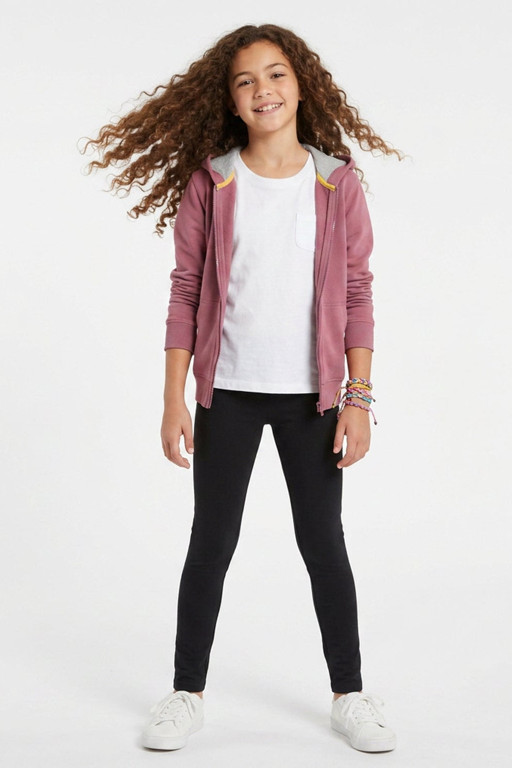 A young girl with long curly brown hair stands against a white studio background wearing a dusty rose zip-up hoodie with yellow accents, a white t-shirt, black skinny pants, and white sneakers.