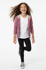 A young girl with curly brown hair is smiling and running in a playful pose, wearing a dusty rose zip-up hoodie with yellow and white striped hood lining, a white t-shirt, black leggings, and black sneakers with white laces, set against a plain white studio background.
