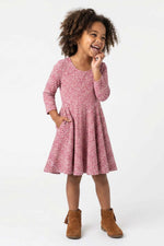 A young girl with curly brown hair wearing a pink and red floral print long-sleeve dress and brown suede ankle boots, standing against a plain white studio background.