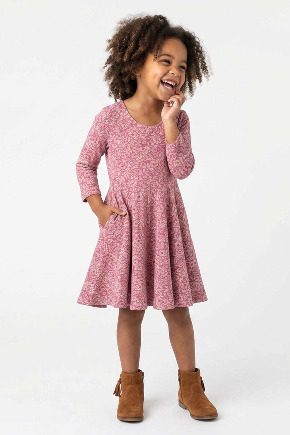 A young girl with curly brown hair wearing a pink and red floral print long-sleeve dress and brown suede ankle boots, standing against a plain white studio background.