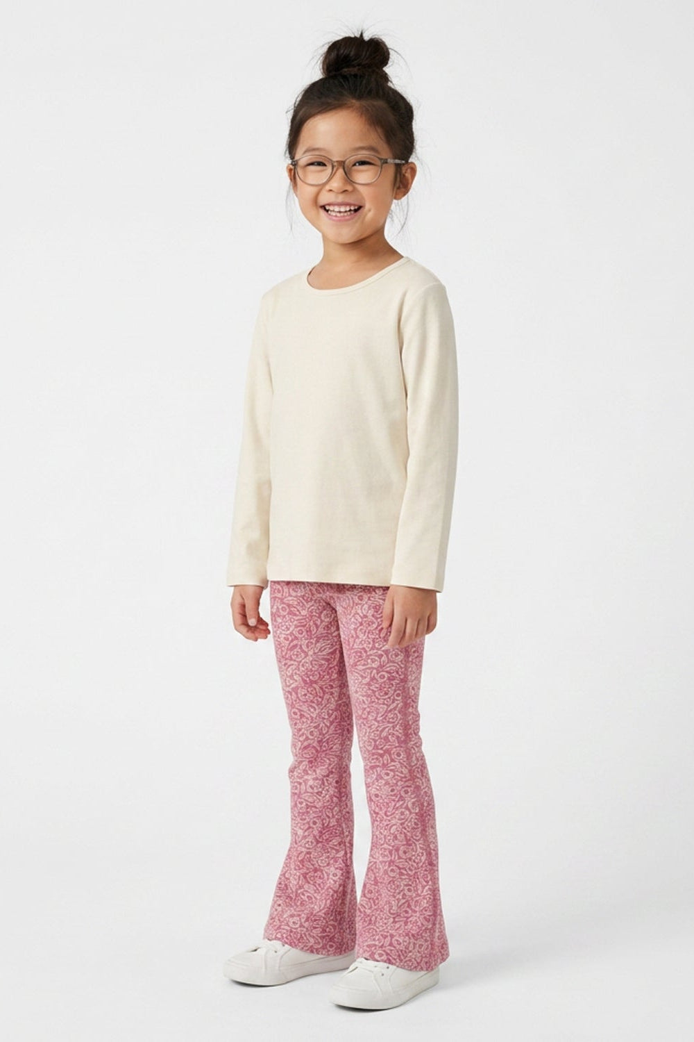 A young girl with glasses and her hair in a bun stands against a white studio background, wearing a cream long-sleeve top, pink patterned flared pants, and white sneakers.
