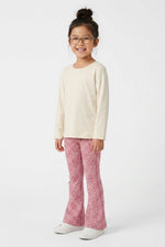 A young girl with glasses and her hair in a bun stands against a white studio background, wearing a cream long-sleeve top, pink patterned flared pants, and white sneakers.
