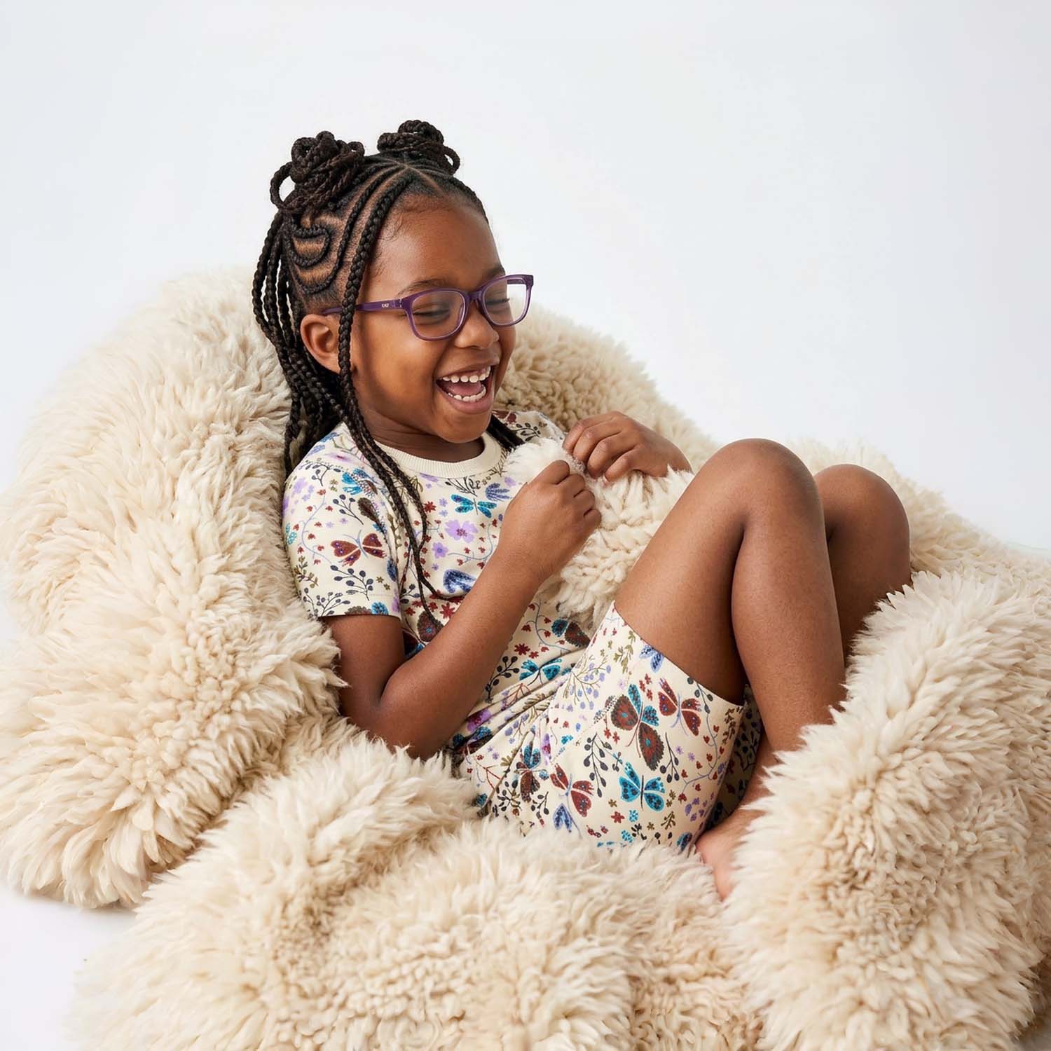 Young girl with braided hair wearing pajamas and glasses, sitting on a fluffy chair.