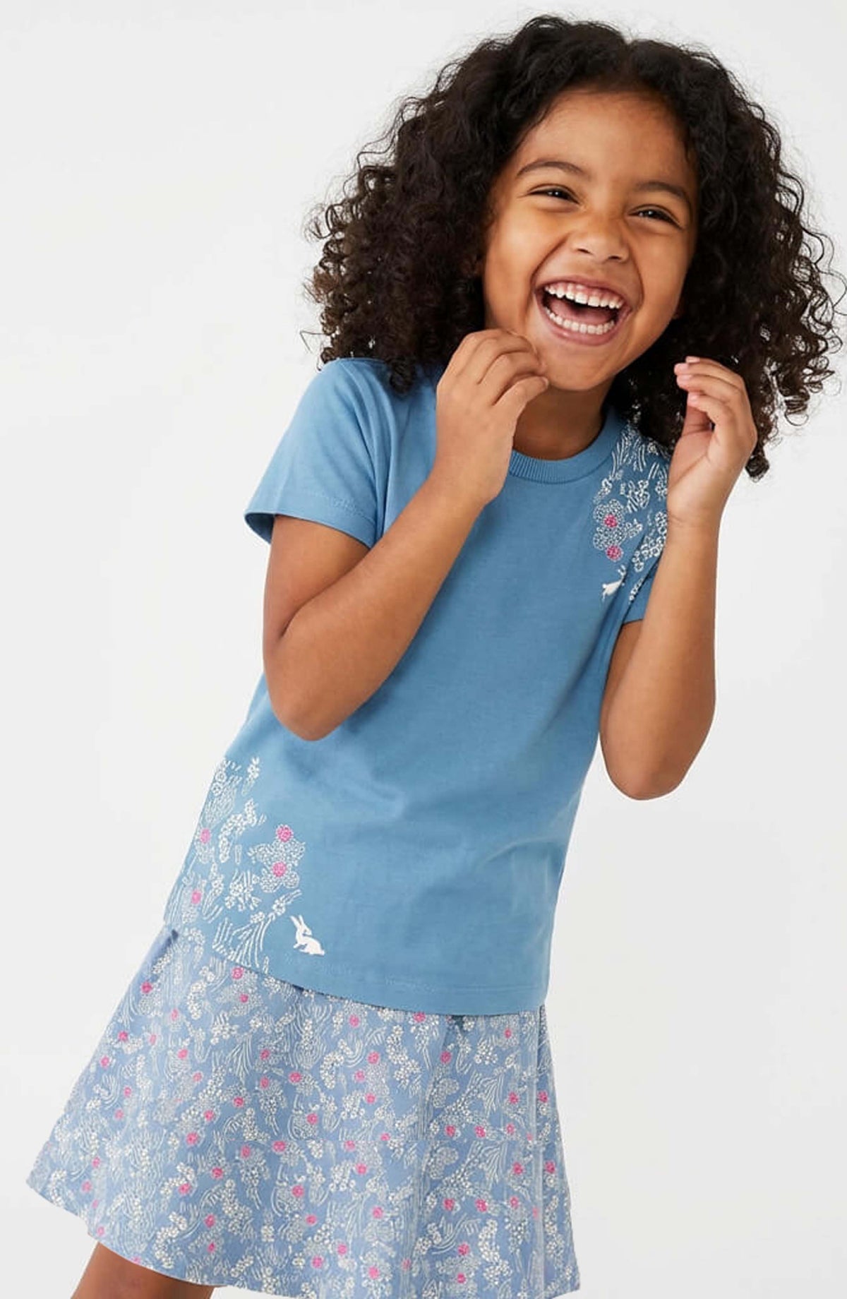 A young girl with curly dark hair is smiling and laughing while wearing a light blue short-sleeve t-shirt with a floral patterned skort, photographed against a clean white studio background.
