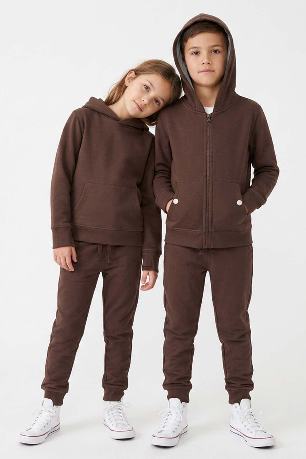 A child's matching two-piece tracksuit in dark brown, shown on two models (a boy and a girl) against a plain white studio background. The set includes a zip-up hoodie with a front pocket and matching jogger pants.