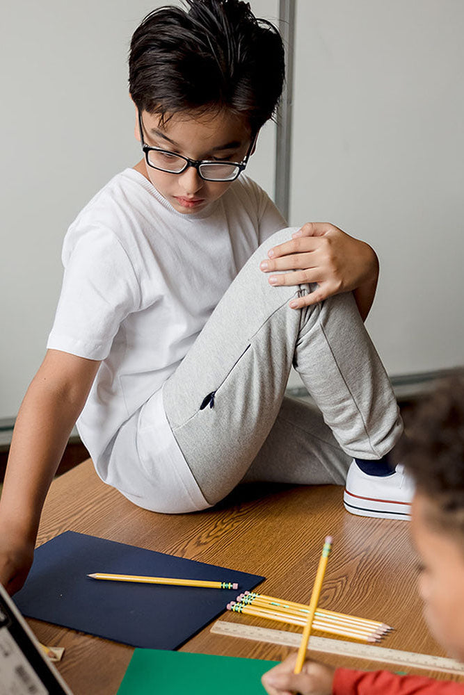 A child wearing a white t-shirt and light gray sweatpants sits on a wooden desk, looking down at a notebook. Another child is visible in the foreground holding a pencil.