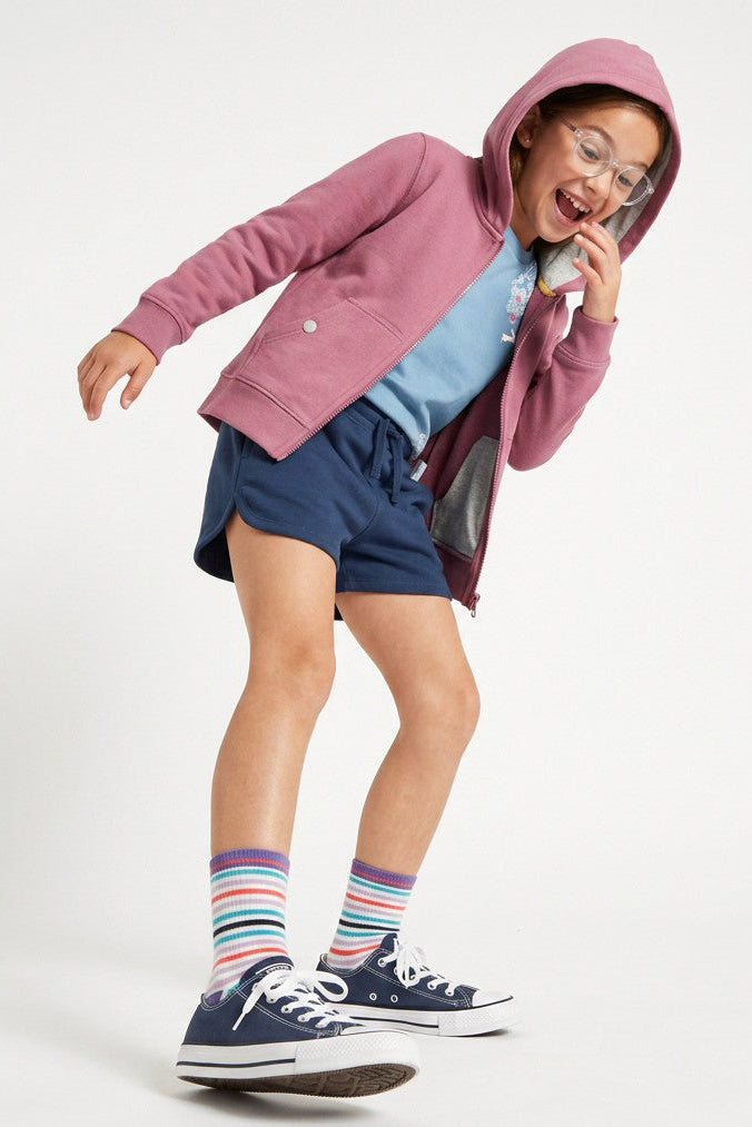 A child modeling a dusty pink zip-up hoodie over a light blue t-shirt, navy shorts, and navy sneakers with colorful striped socks, posing on a white studio background.