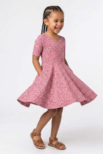 A young girl with braided hair wearing a pink short-sleeve dress with a white floral pattern and brown sandals, standing against a plain white studio background.