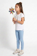 A young girl with her hair in a bun stands against a white studio background, wearing a white t-shirt, light blue skinny jeans, and white sneakers, while holding a colorful pinwheel.