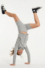 A person performing a handstand in a light gray long-sleeve crop top and matching shorts, with black high-top sneakers, against a plain white studio background.