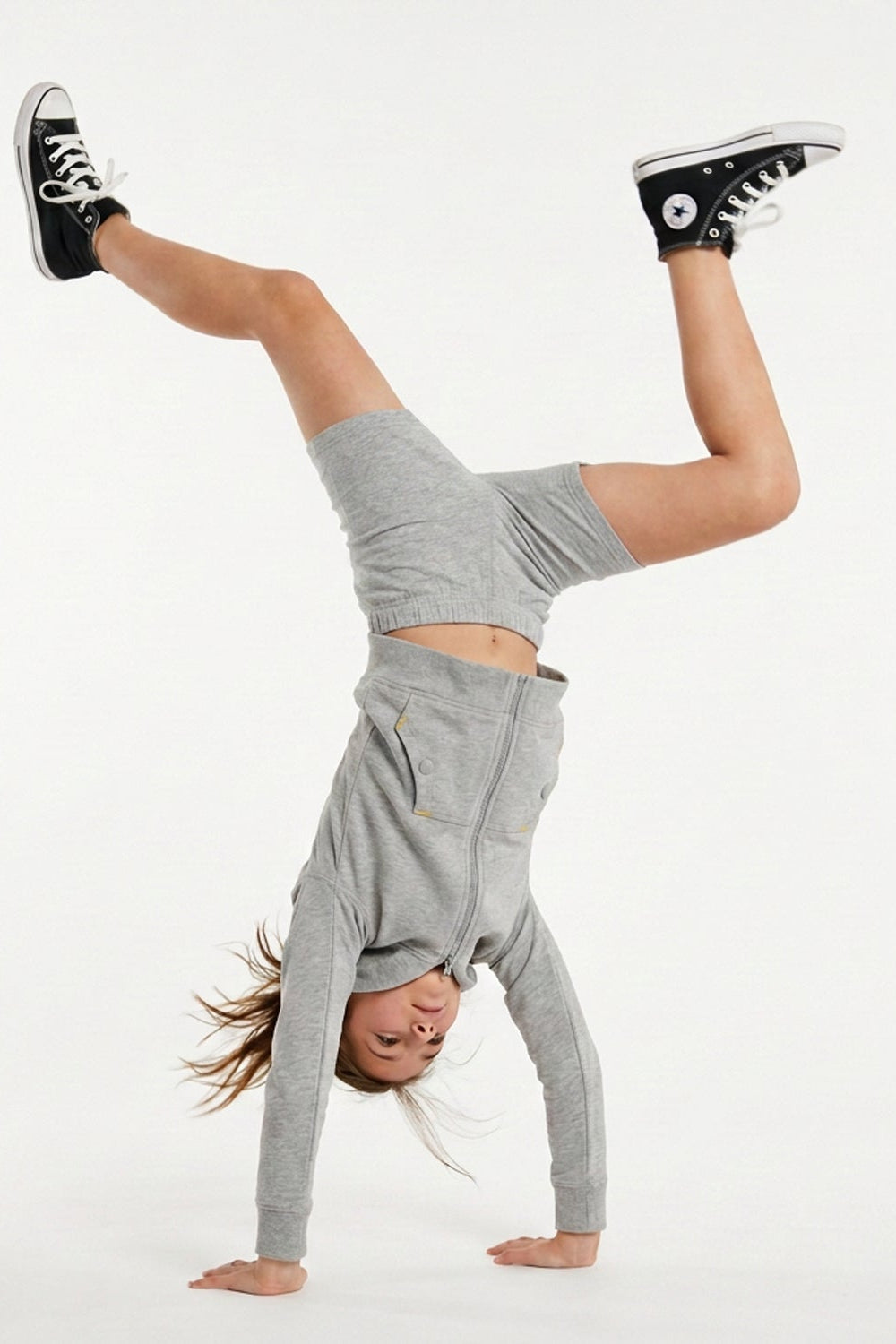 A person performing a handstand in a light gray long-sleeve crop top and matching shorts, with black high-top sneakers, against a plain white studio background.