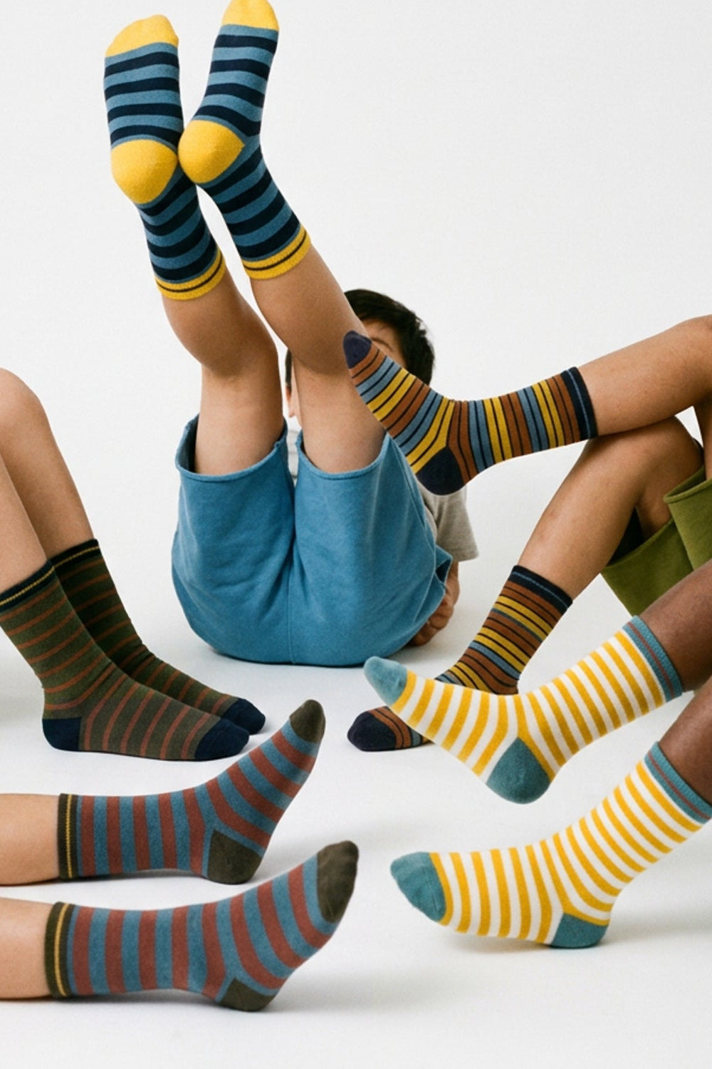 Several children sitting on a white floor wearing colorful striped socks. The socks feature patterns of navy, yellow, blue, green, and brown stripes with solid yellow toes and heels.
