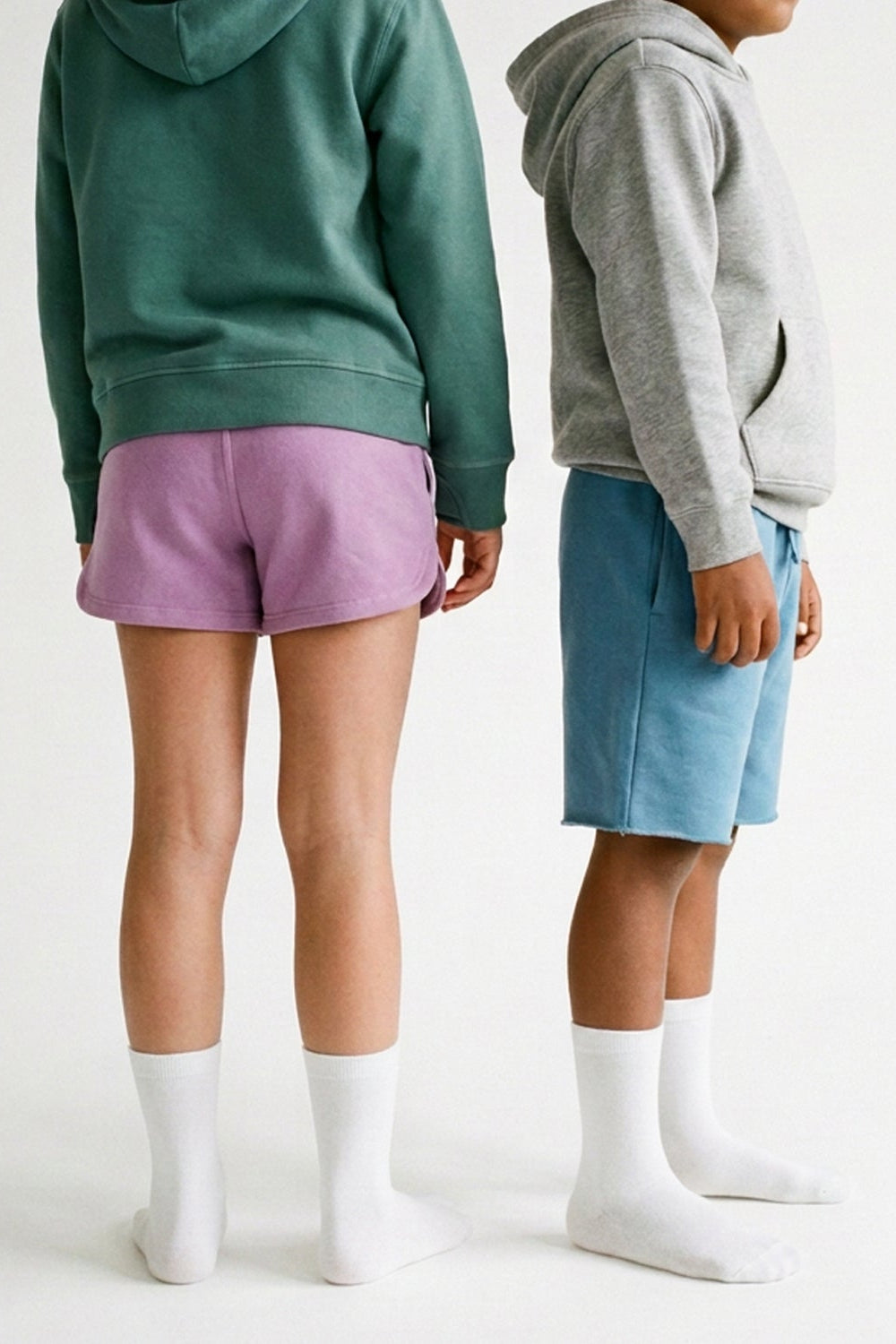 Two children standing in profile wearing matching hoodies and shorts, with one in a dark green hoodie and lavender shorts and the other in a heather gray hoodie and light blue shorts. Both wear white socks against a plain white studio background.
